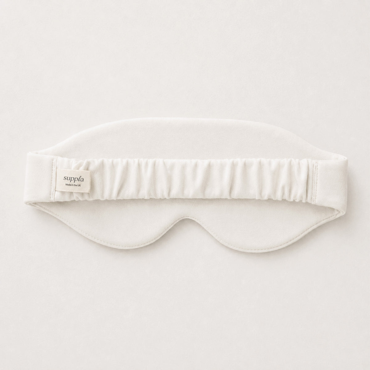 A white sleep mask with an elasticized top band, featuring a small fabric label that reads 'suppla'. The mask is laid flat against a clean, off-white background.