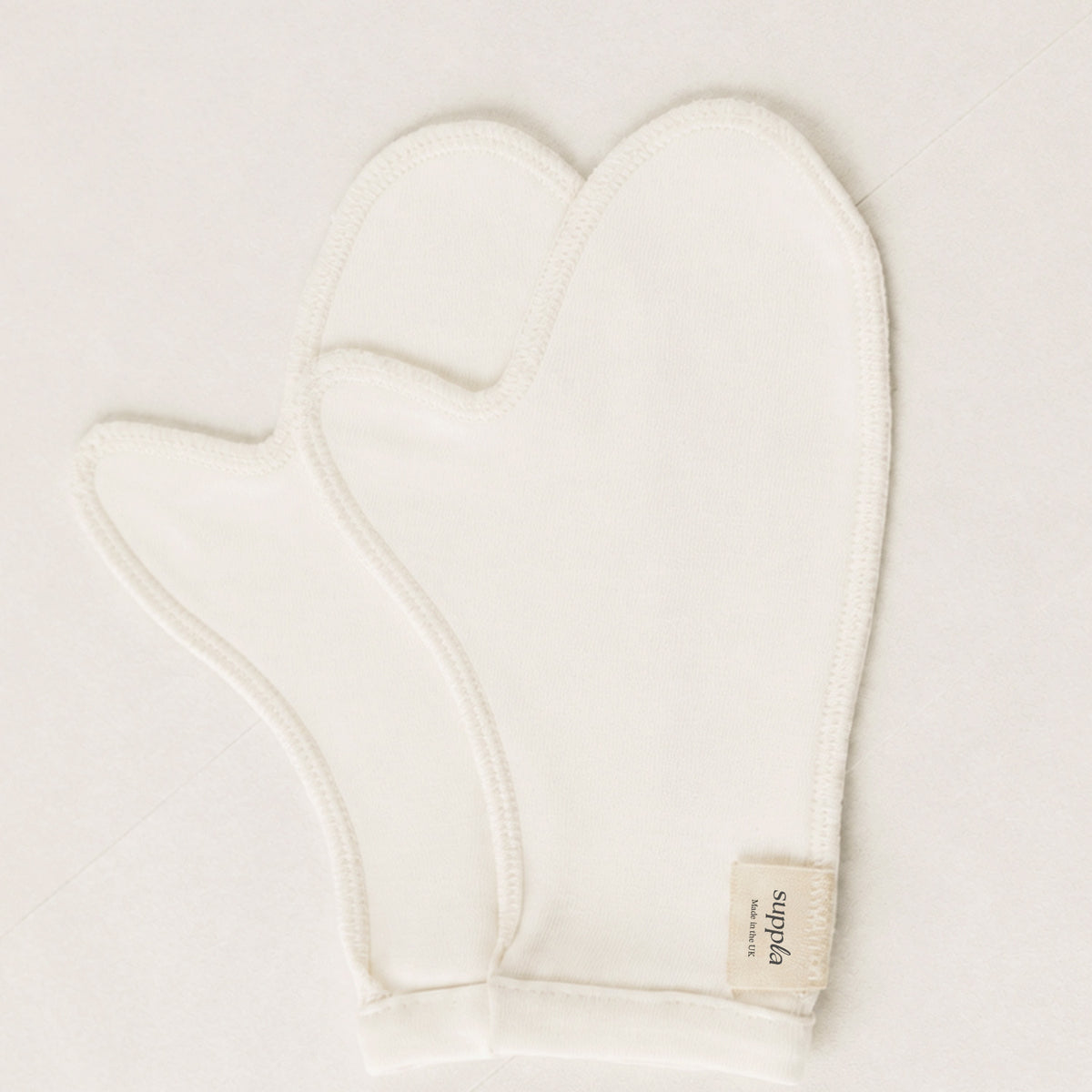 A pair of off-white cotton cleaning mittens with a small beige fabric brand tag reading 'suppla' on a matching off-white background.