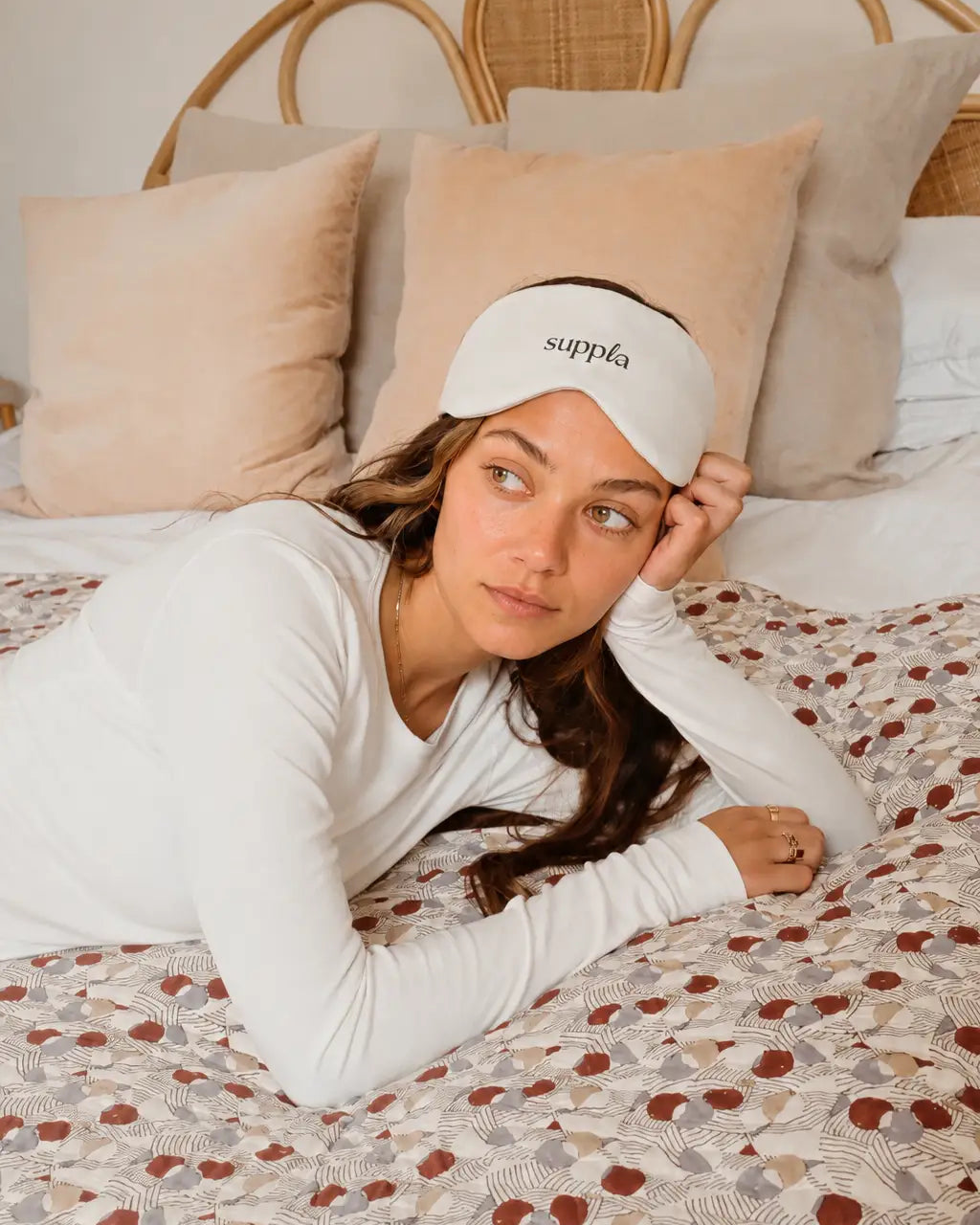 A woman lies on a bed wearing a white long-sleeve top and a white sleep mask with the brand name 'suppla' printed on it. The bed has beige and brown patterned bedding, and a rattan headboard is visible in the background.
