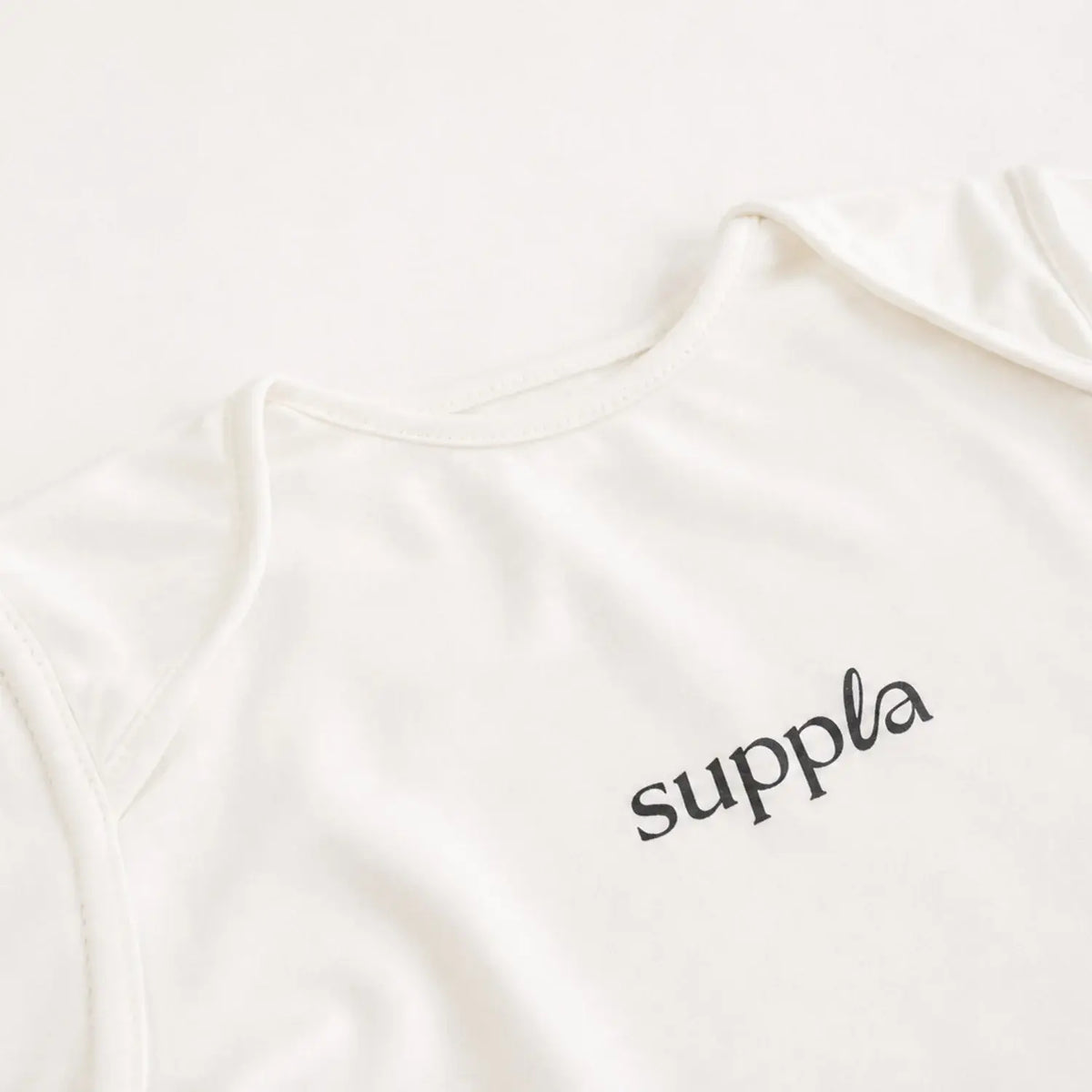 Close-up of a cream-colored  sleepsuit with the brand name 'suppla' printed in black script on the chest, set against a matching cream background.