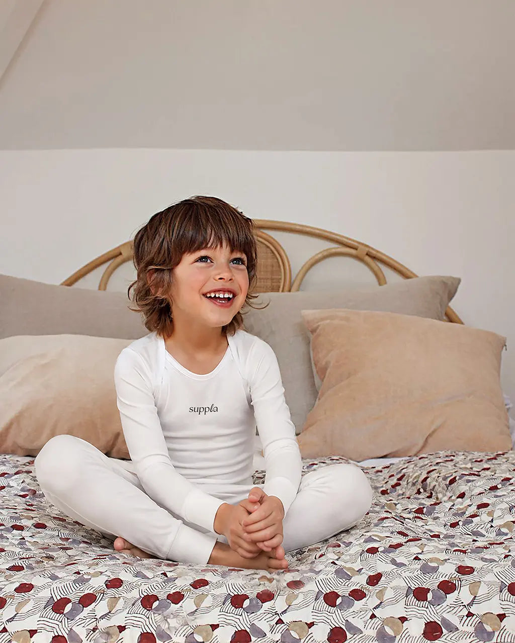 A young child with brown hair sits cross-legged on a patterned bedspread, wearing a sleepsuit. The top features the brand name 'suppla' printed in black on the chest.