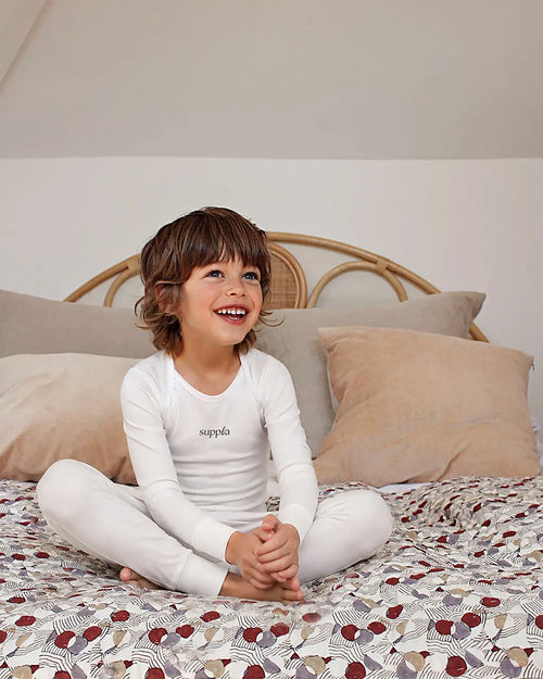 A young child with brown hair sits cross-legged on a patterned bedspread, wearing a sleepsuit. The top features the brand name 'suppla' printed in black on the chest.