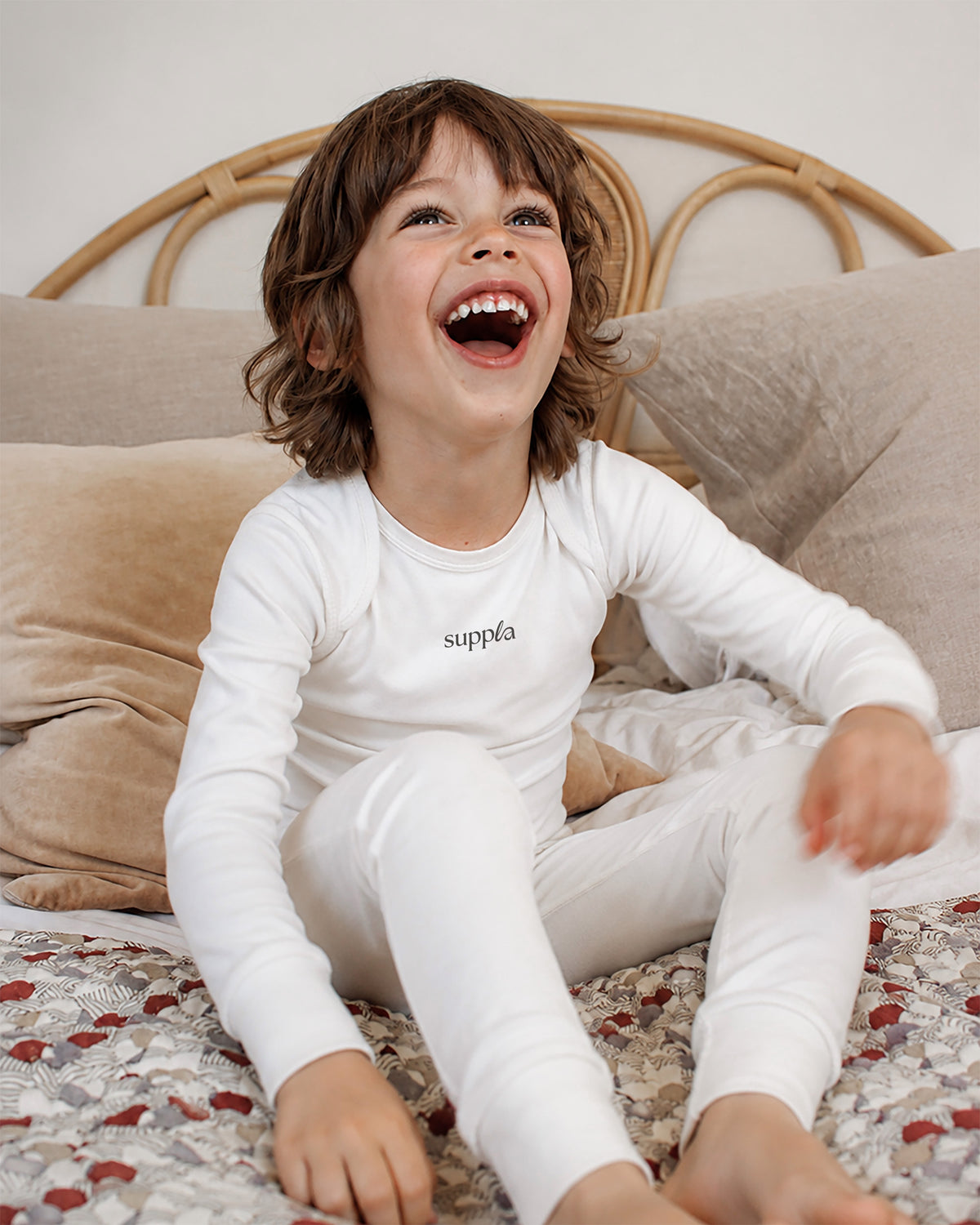 A young child with brown hair sits smiling on a bed wearing a sleepsuit. The shirt has the brand name 'suppla' printed in black on the chest.