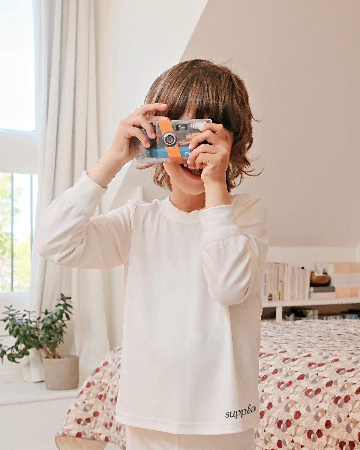 A child wearing a white long-sleeve pajama top with 'suppla' printed near the hem, holding a toy camera in front of their face in a bright bedroom setting.
