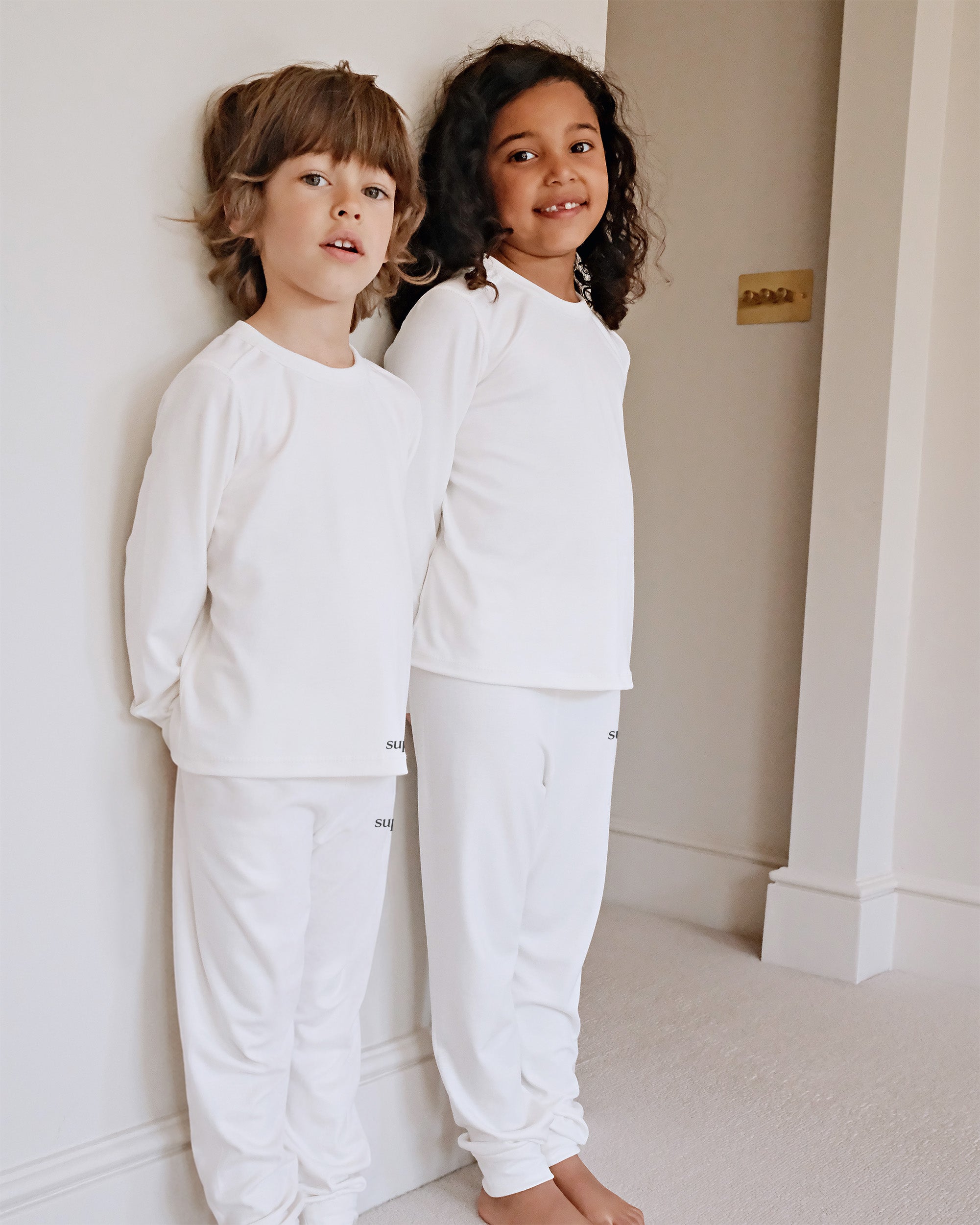 Two children standing barefoot indoors against a white wall, wearing matching white long-sleeve tops and white pants.