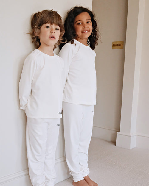 Two children standing barefoot indoors against a white wall, wearing matching white long-sleeve tops and white pants.