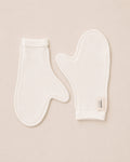 Two white cotton baby mittens laid flat on a light beige background, with one mitten showing a small fabric brand tag that reads Suppla