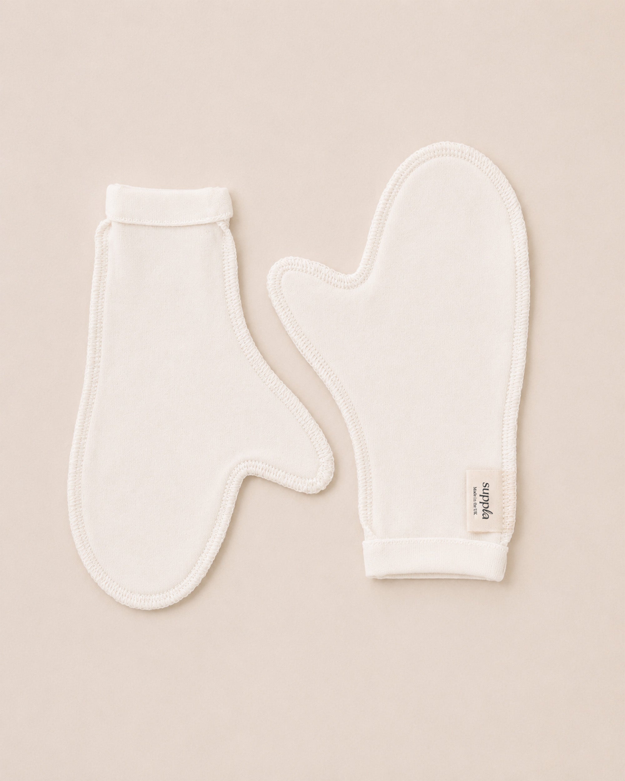 Two white cotton baby mittens laid flat on a light beige background, with one mitten showing a small fabric brand tag that reads Suppla