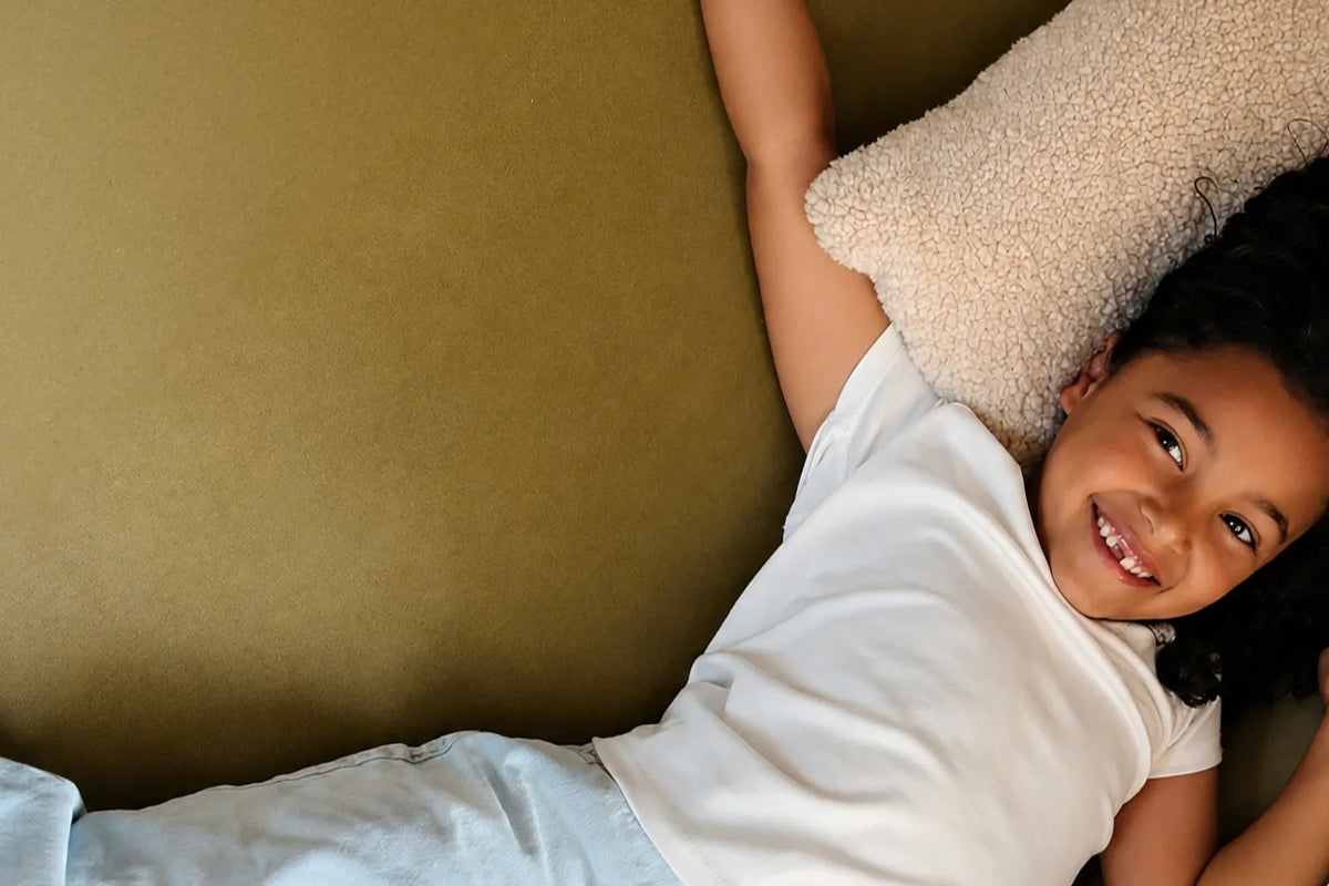 A smiling young girl with dark curly hair lies on her back on a textured olive green sofa, resting her head on a cream textured pillow and wearing a white t-shirt and light blue jeans.