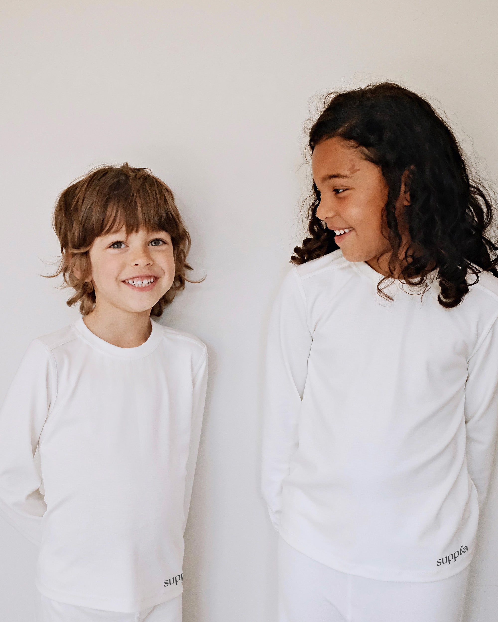 Two children, a boy and a girl, smiling and looking at each other while wearing matching white long-sleeve shirts and white pants with the brand name 'suppla' printed near the hem.