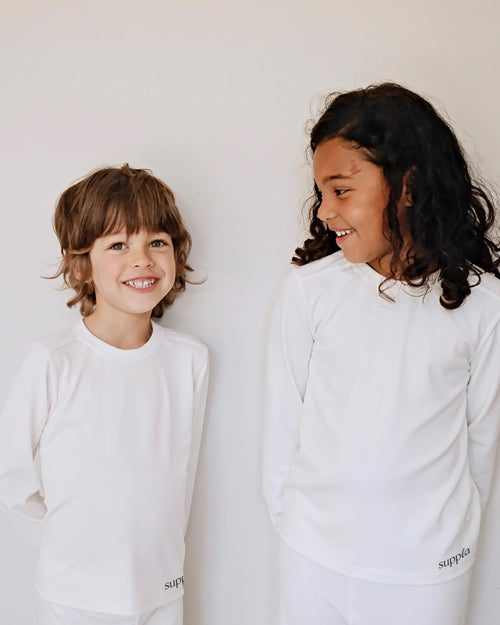 Two children, a boy and a girl, smiling and looking at each other while wearing matching white long-sleeve shirts and white pants with the brand name 'suppla' printed near the hem.
