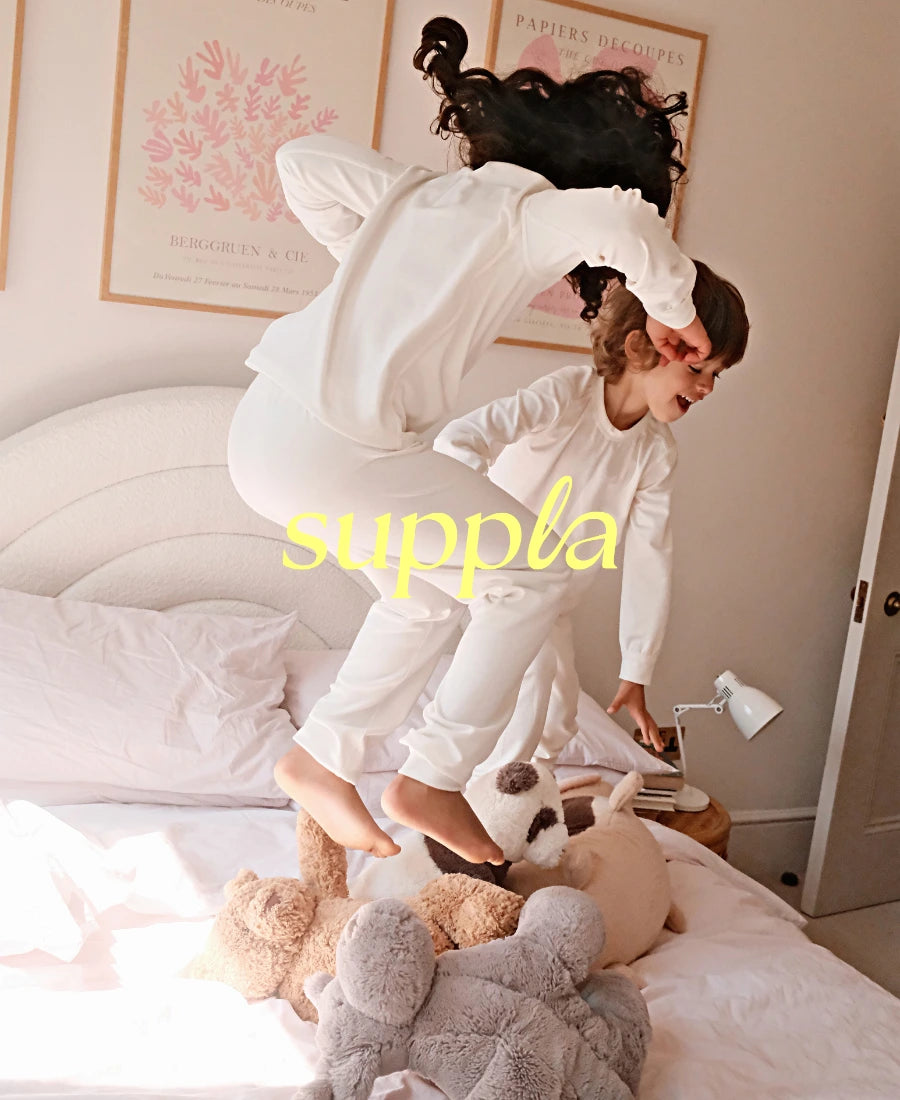 Two children playing on a bed with stuffed animals, wearing white outfits, in a room with 'suppla' branding.