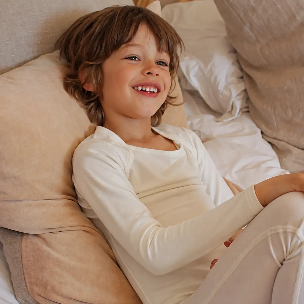 Child lying on a bed wearing white suppla calm therapeutic eczema wear, surrounded by beige pillows.
