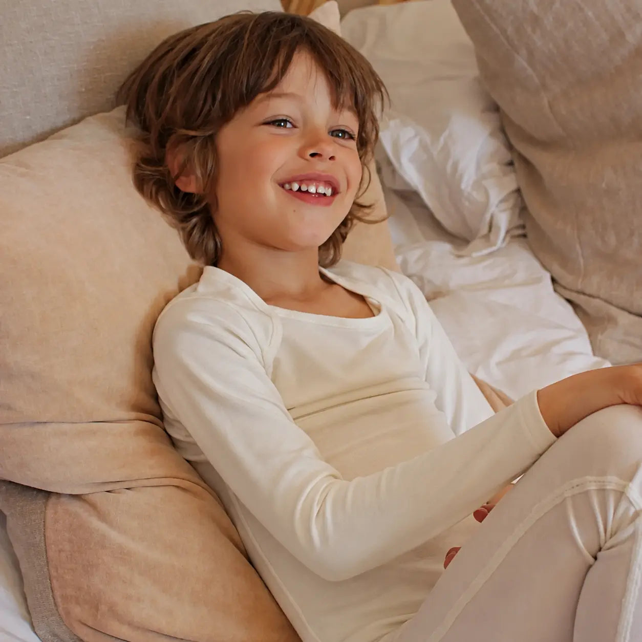 Child lying on a bed wearing white suppla calm therapeutic eczema wear, surrounded by beige pillows.