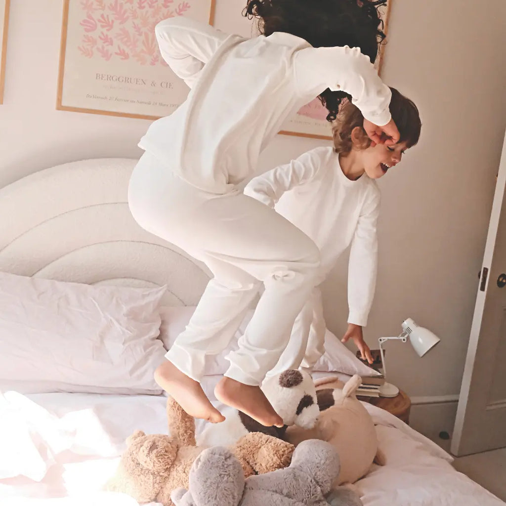 Two children in Suppla Core eczema pajamas bouncing on a bed with stuffed animals.