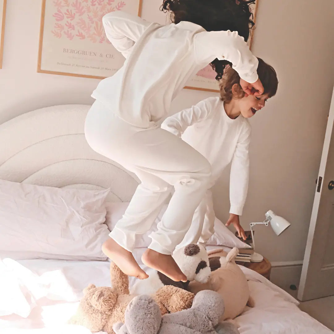 Two children in Suppla Core eczema pajamas bouncing on a bed with stuffed animals.