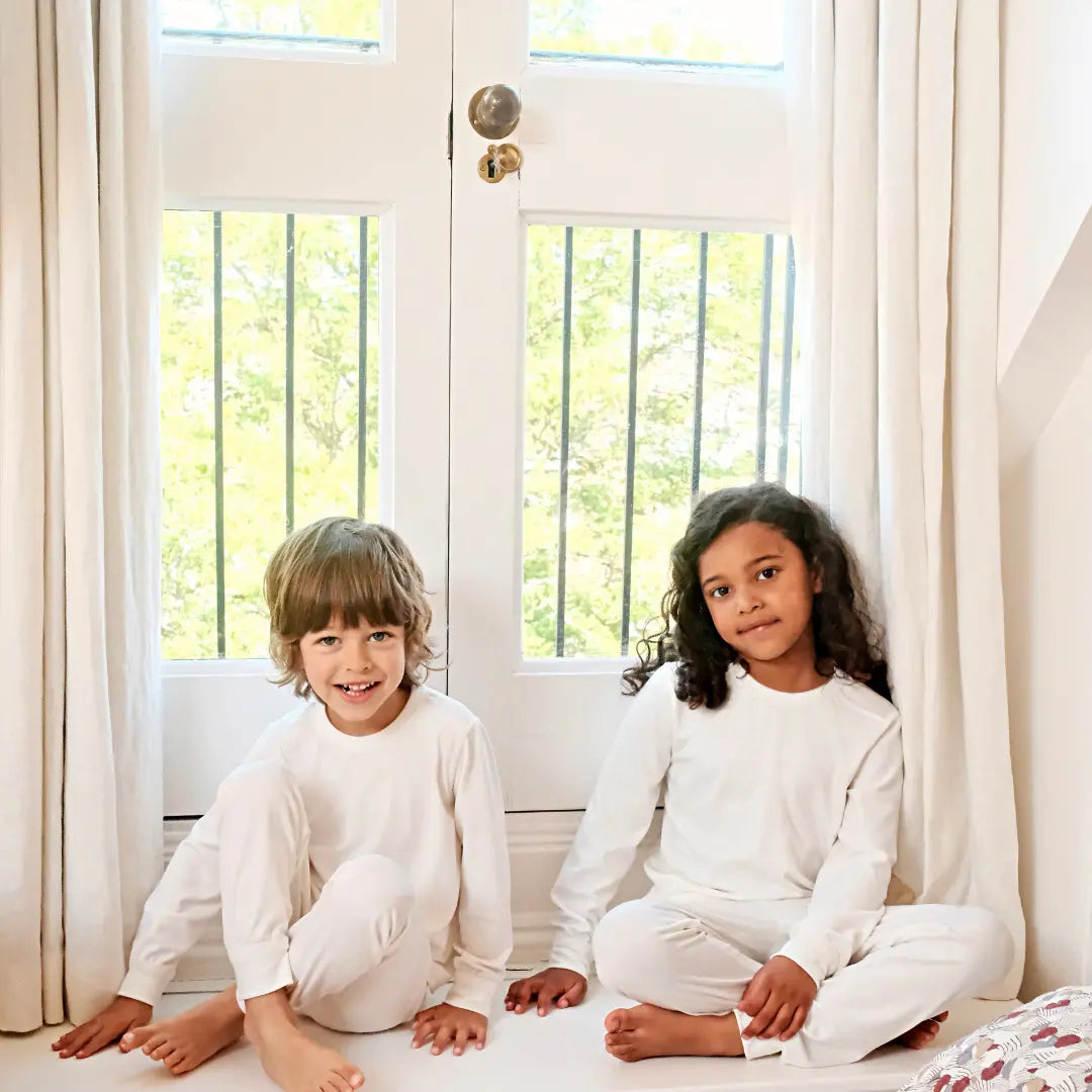 Two children in white Suppla eczema pajamas sitting on a bed in a bright room with large windows.