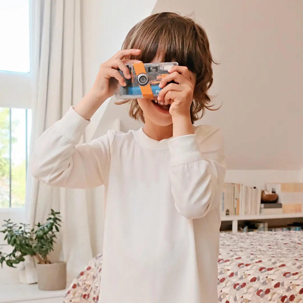 Child wearing white suppla eczema wear holding a toy camera in a bright room with plants and furniture.